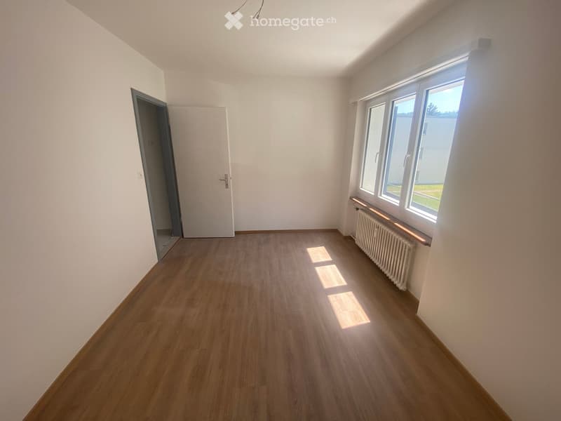 Empty room with wooden flooring, white walls, and large windows allowing natural light. Features a radiator under the windows and a door leading to another space.