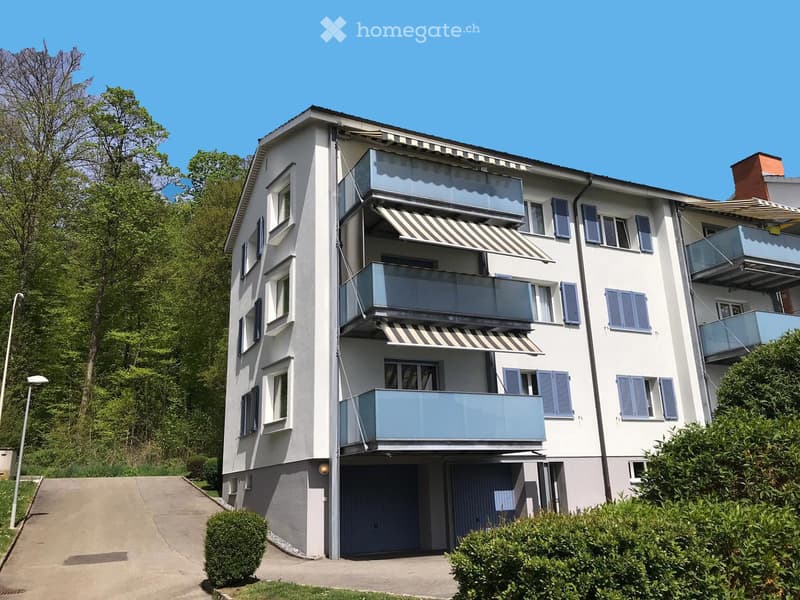 Modern three-story residential building with white facade, multiple balconies, striped awnings, and blue shutters. Features a garage entrance and is surrounded by greenery.