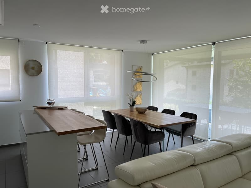 Modern living and dining area with large windows covered by roller blinds, a white leather sofa, a wooden dining table with six chairs, and a kitchen island with bar stools.