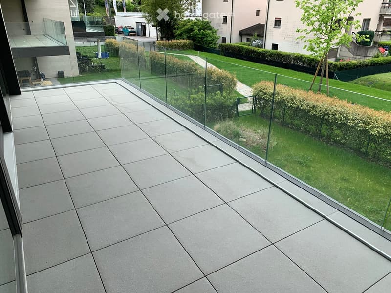 Modern balcony with large glass railing, tiled floor, and view of a well-maintained garden with green grass, bushes, and trees.