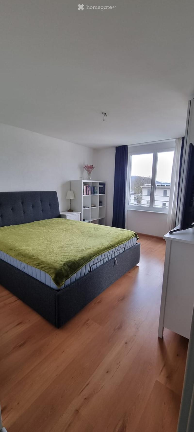 large double bed with green blanket, white headboard, wooden floor, white walls, white shelving unit, lamp, TV on table