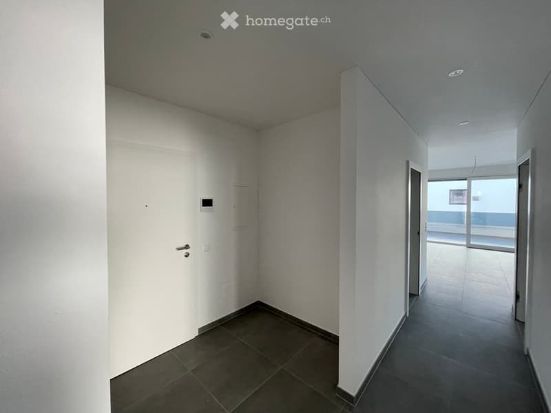 Empty hallway with white walls, dark tiled floor, and recessed lighting. A door is visible on the left, and a glass door at the end leads to another room.