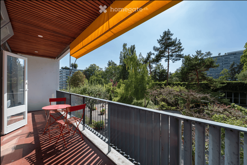 Balcony with red table and chairs, wooden ceiling, metal railing, and a view of greenery and trees.