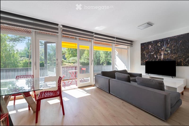Modern living room with large windows and sliding doors leading to a balcony, featuring a grey sectional sofa, a glass dining table with red chairs, and a television on a white stand.