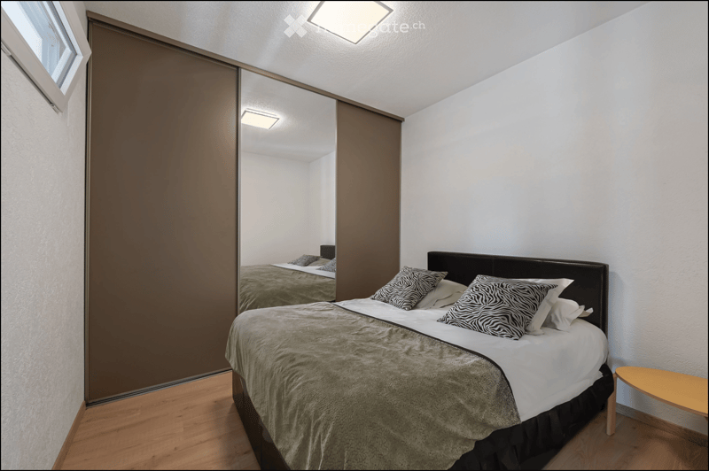 Modern bedroom with a double bed, nightstand, mirror, and small table. The room features light-colored walls, wooden flooring, and a large mirror reflecting the bed.