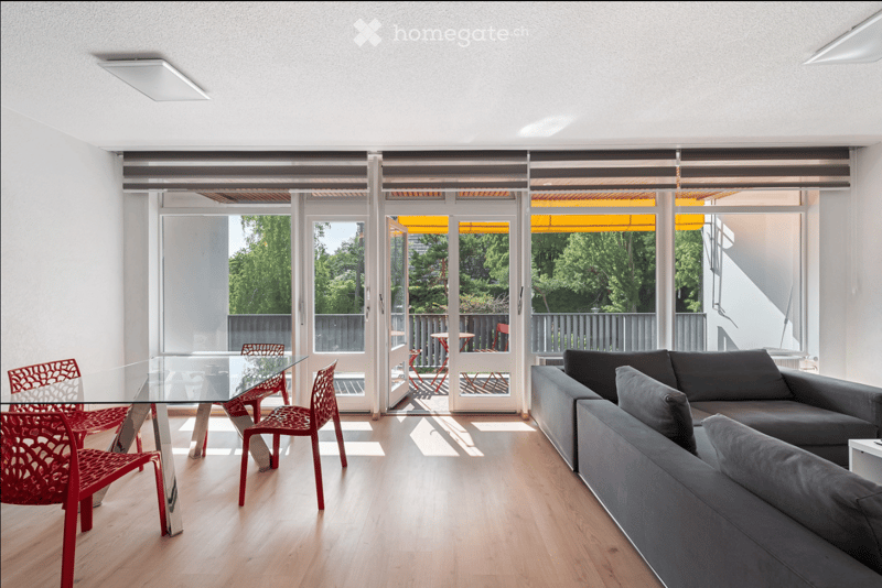 Modern living and dining area with light wood flooring, large glass doors leading to a balcony, grey sofa, and a glass dining table with red chairs.