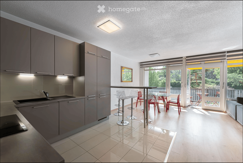 Modern kitchen with grey cabinets, a sink, and a dining area with red chairs and a glass table. Large windows and a sliding door lead to a balcony.