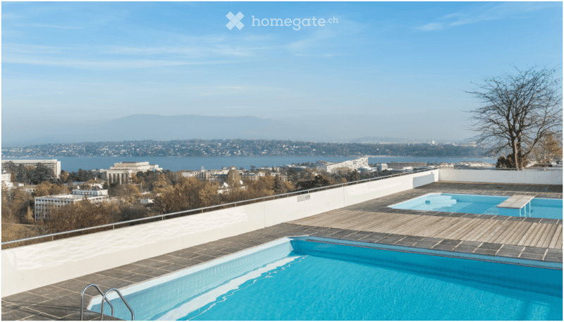 Rooftop terrace with a swimming pool, overlooking a cityscape and water body with distant mountains.