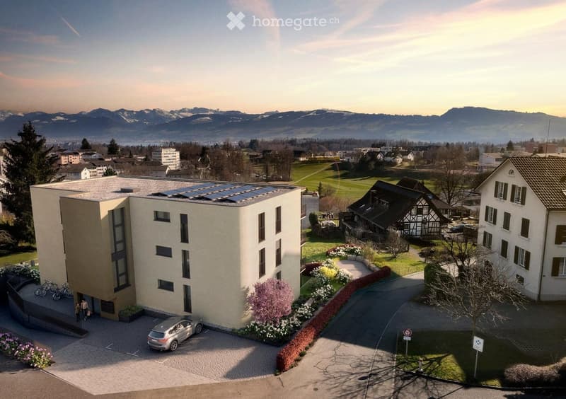 A modern multi-story building with solar panels on the roof, surrounded by a landscaped garden with flowers and trees. A silver car is parked in the driveway, and bicycles are near the entrance. The background shows a scenic view of mountains and a town.