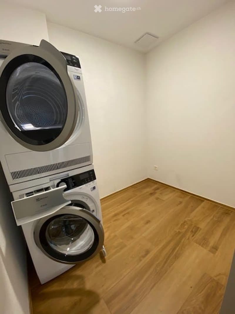 A room with a stacked washer and dryer, hardwood floor, white walls, and power outlet