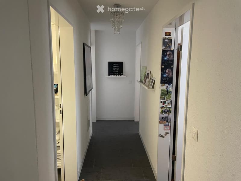 A clean, well-lit hallway with white walls, dark tiled flooring, and a chandelier hanging from the ceiling. There are open doors on both sides leading to other rooms, and small shelves with framed pictures and a motivational sign are visible.