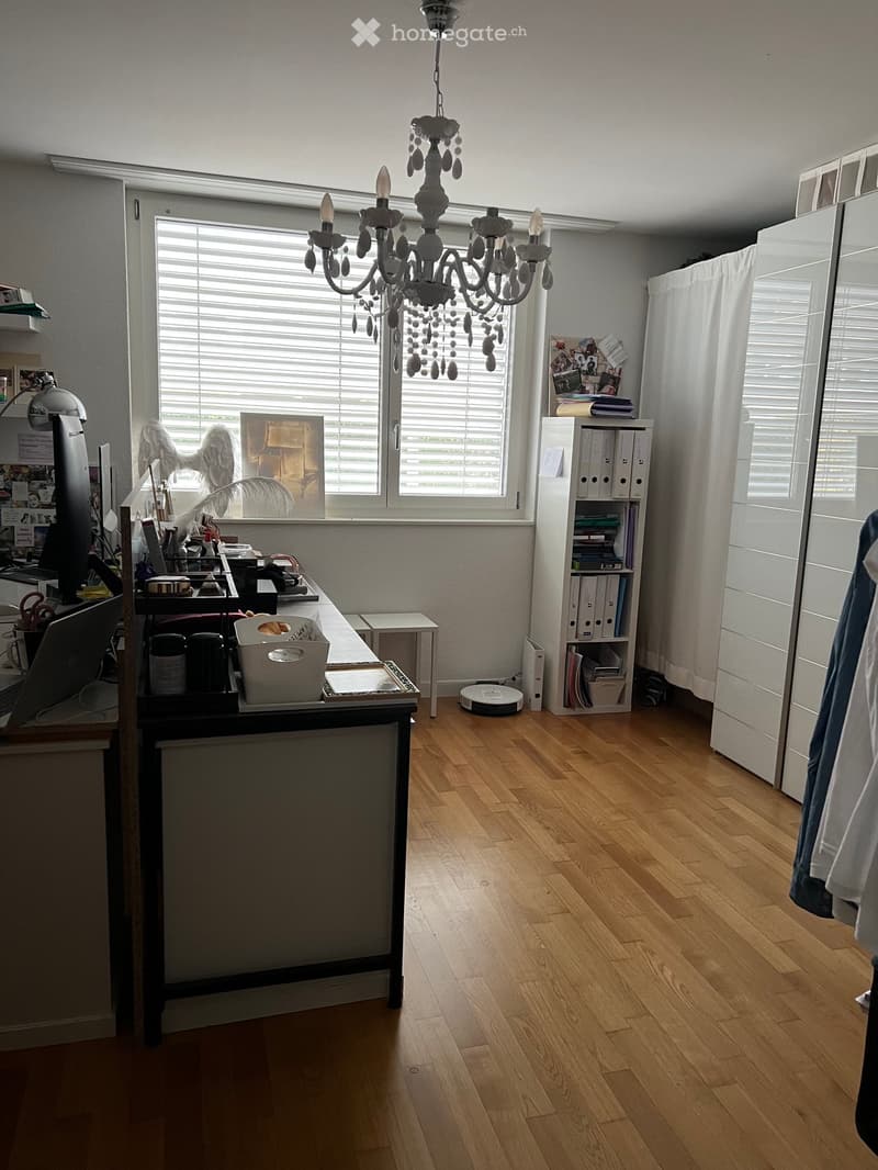 Well-lit room with wooden flooring, large window with blinds, chandelier, desk with office supplies, bookshelf, and a small table.