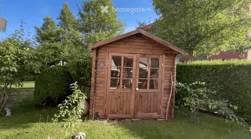 A wooden shed with glass doors is situated in a well-maintained garden, surrounded by greenery and trees.