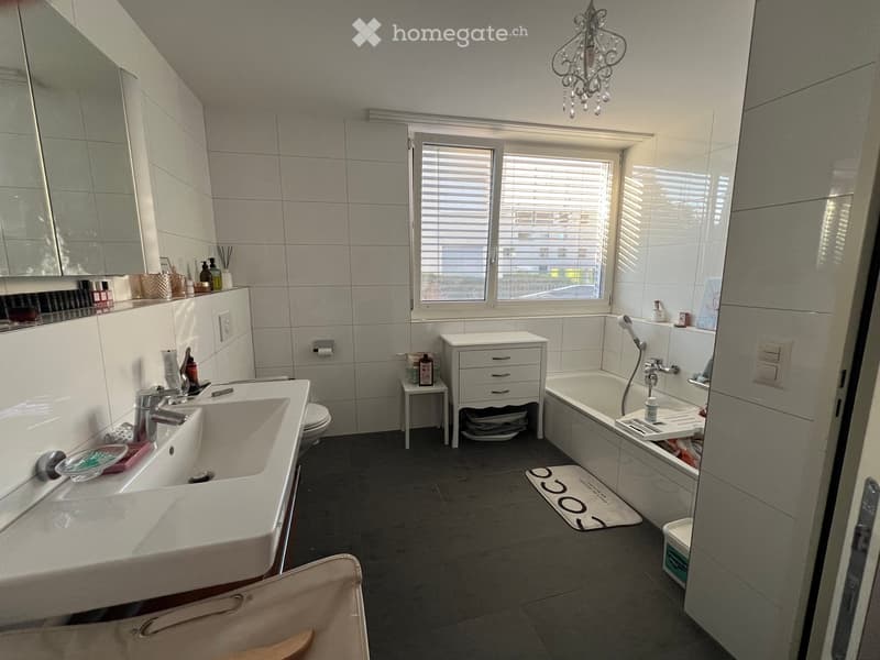Modern bathroom with double sink, bathtub, large mirror, and a small cabinet. Features a chandelier and a window with blinds.