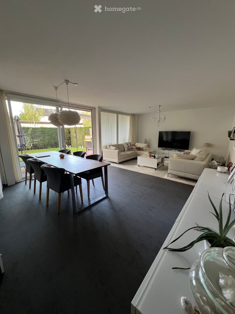 Modern living and dining area with dark flooring, white walls, large windows, and a view of the garden. Features a sectional sofa, coffee table, dining table with chairs, and a television on a stand.