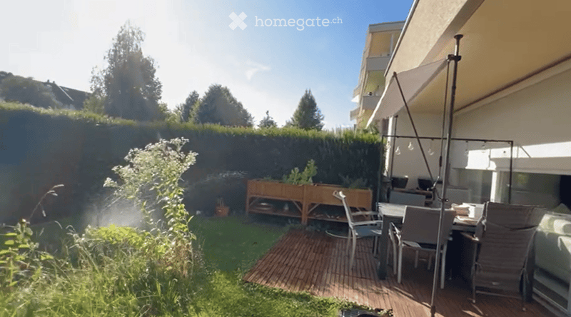 A well-maintained garden with a wooden deck, outdoor dining set, and a raised planter box. The area is enclosed by a hedge and features a modern awning attached to the building.