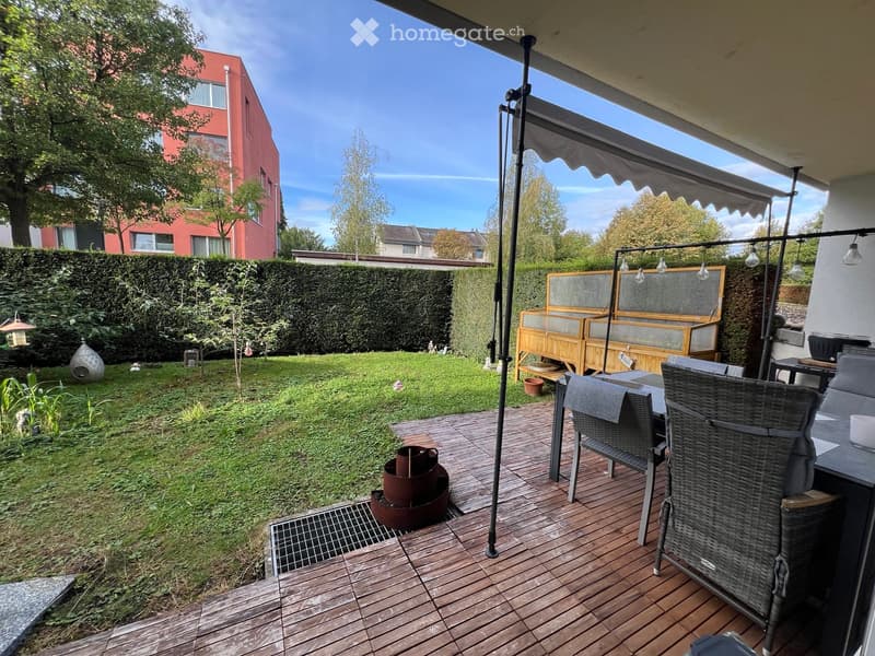 Wooden deck with outdoor furniture, including wicker chairs and a table, surrounded by a well-maintained lawn and hedge. Features a fire pit, a covered seating area, and a view of neighboring buildings.