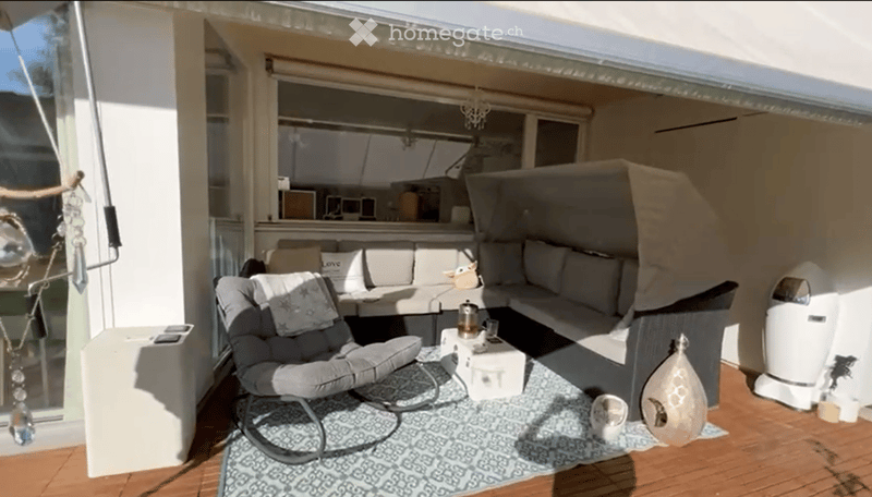 A cozy, furnished balcony with a grey sofa, a single armchair, a small coffee table, and decorative items including a vase and a table lamp. The space features wooden flooring and a patterned rug.