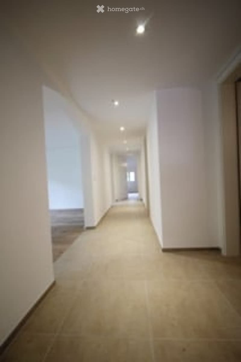 Empty hallway with white walls, beige tiled floor, and recessed lighting. No furniture or items visible.