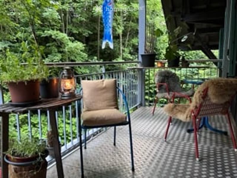 A cozy balcony with a wooden table, potted plants, and two chairs, one with a fur throw, overlooking a lush green forest.