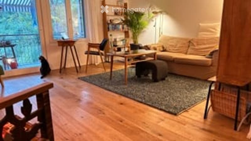 Bright living room with wooden flooring, large windows offering a view, a cream-colored sofa, coffee table, chairs, and a small table with a lamp.