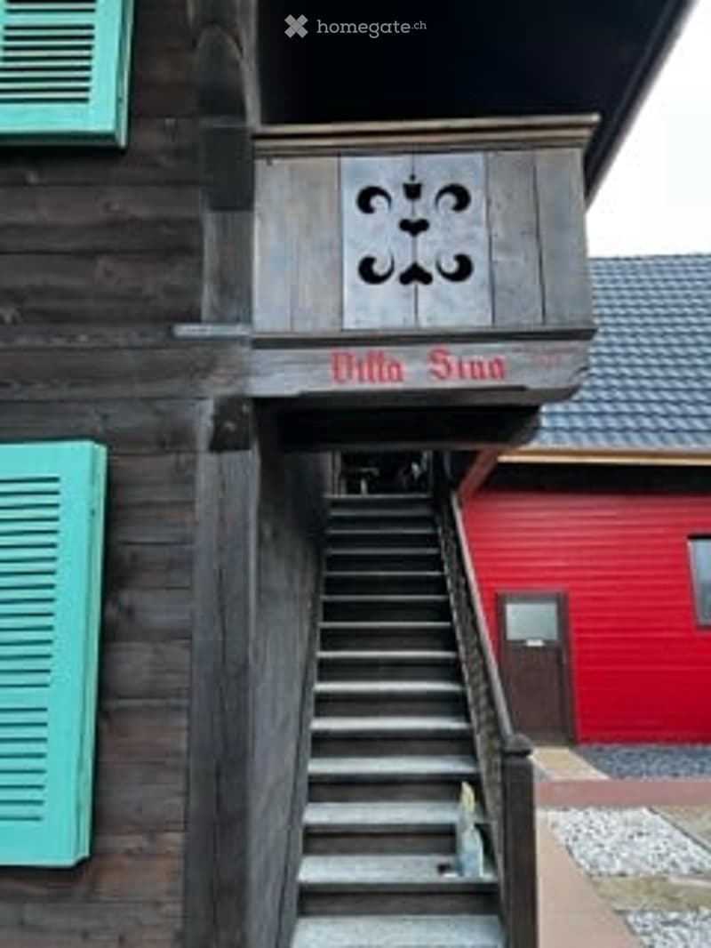 A wooden building with a staircase leading to an upper level, featuring a sign that reads "Villa Sina" and decorative carvings above the entrance.