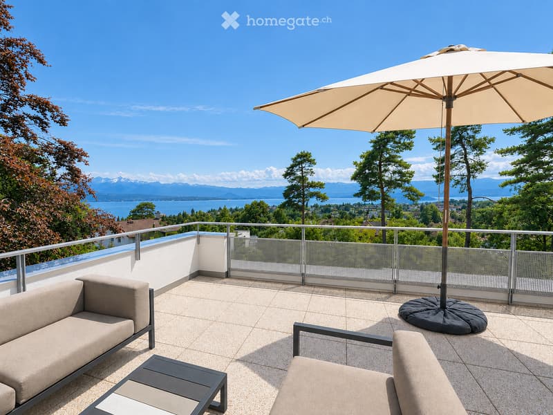 Balcony with a sofa, chair, table, and umbrella, overlooking a scenic lake and mountain view.