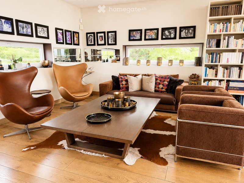 Modern living room with a brown leather sofa, two leather armchairs, a wooden coffee table, a bookshelf, and decorative mirrors on the wall.