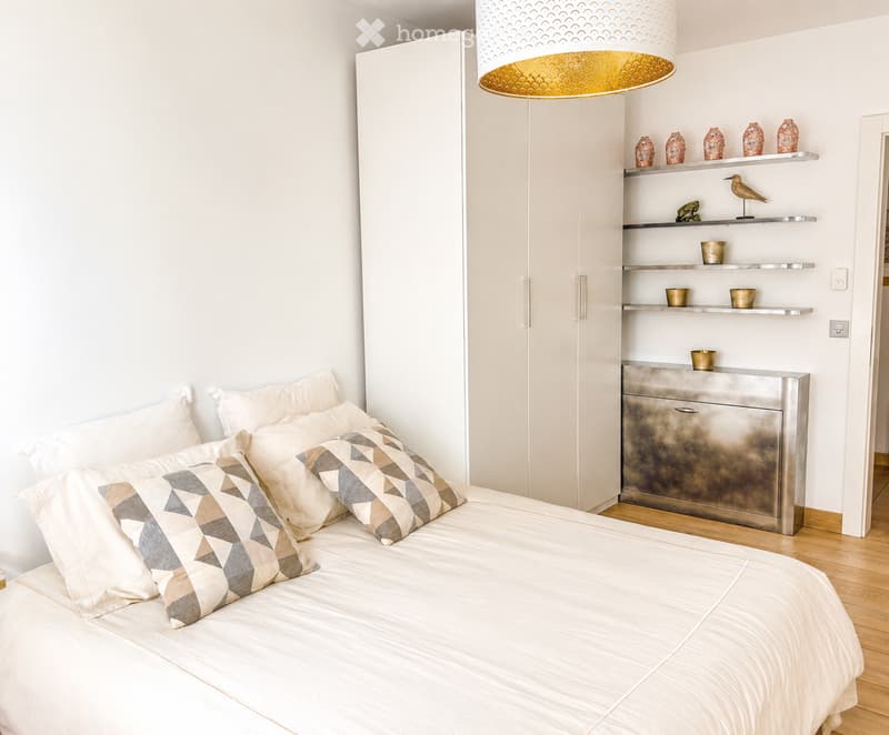 Modern bedroom with a large bed, geometric patterned pillows, white bedding, a tall wardrobe, and decorative shelves with vases and a small bird figurine.