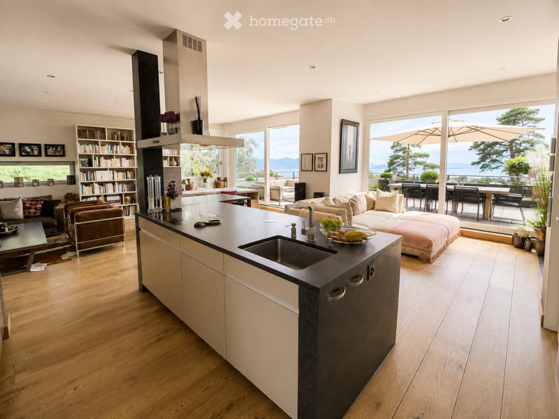 Open-plan living and kitchen area with wooden flooring, large windows offering a scenic view, a central kitchen island with sink and faucet, a sofa, coffee table, bookshelf, and dining area with chairs and table.