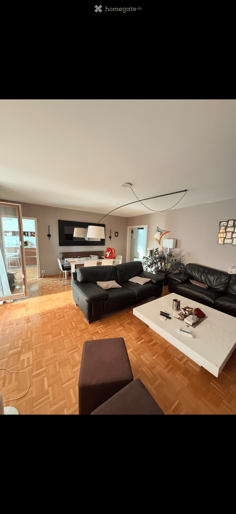 Modern living room with wooden flooring, black leather sofa, white coffee table, ottoman, and a dining area visible through an open doorway.