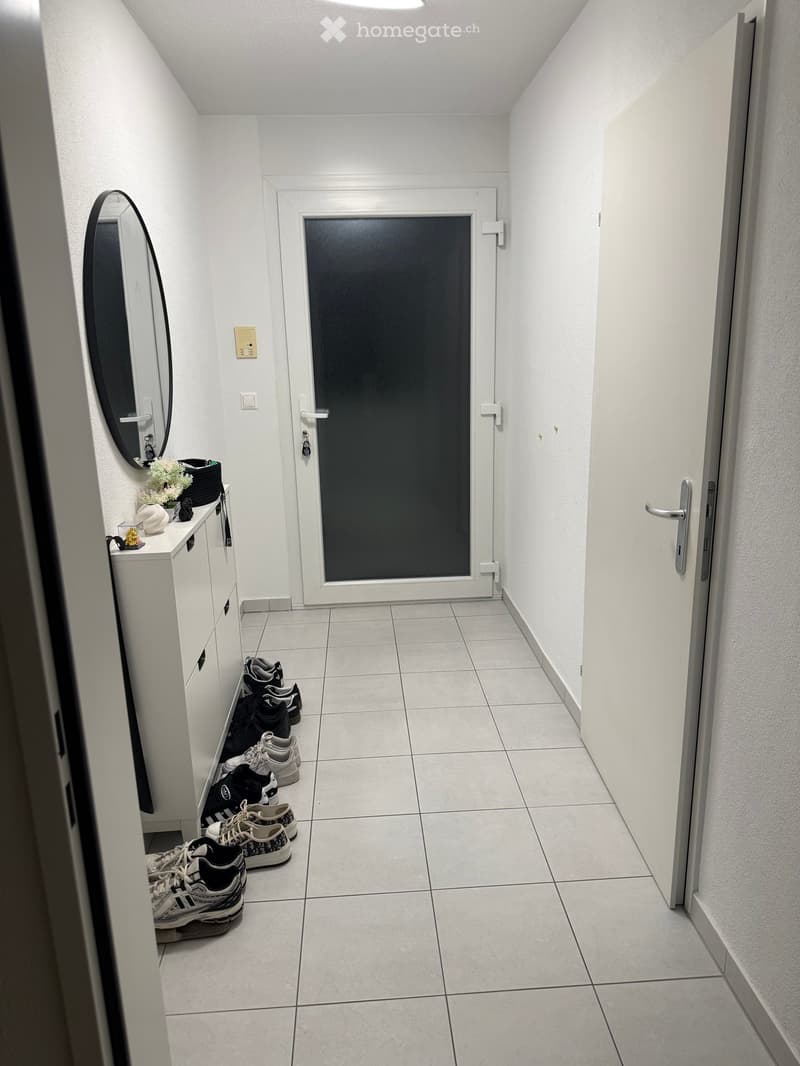 A narrow hallway with white tiled flooring, white walls, and a closed door at the end. A small cabinet with a mirror and decorative items is visible on the left, along with several pairs of shoes placed on the floor.