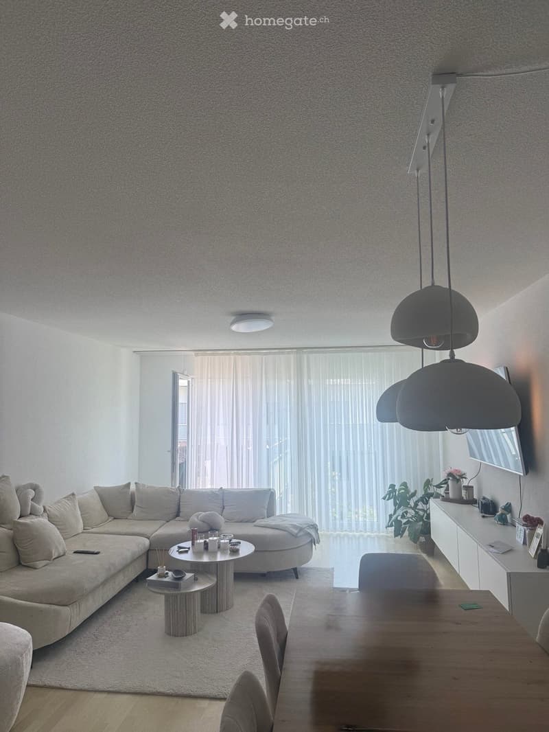 Modern living room with a large sectional sofa, coffee table, dining table, pendant lights, and a TV screen. The space features large windows with sheer curtains and a neutral color palette.