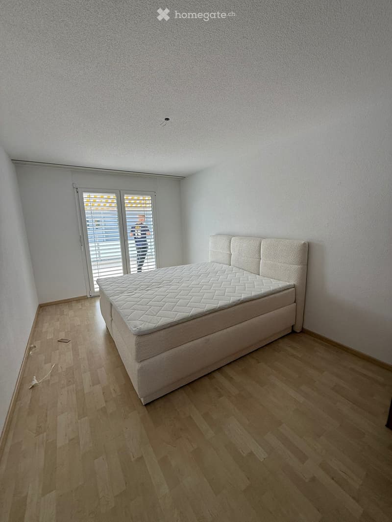 Minimalist bedroom with a single bed, headboard, and large window with blinds. Light wooden flooring and white walls.