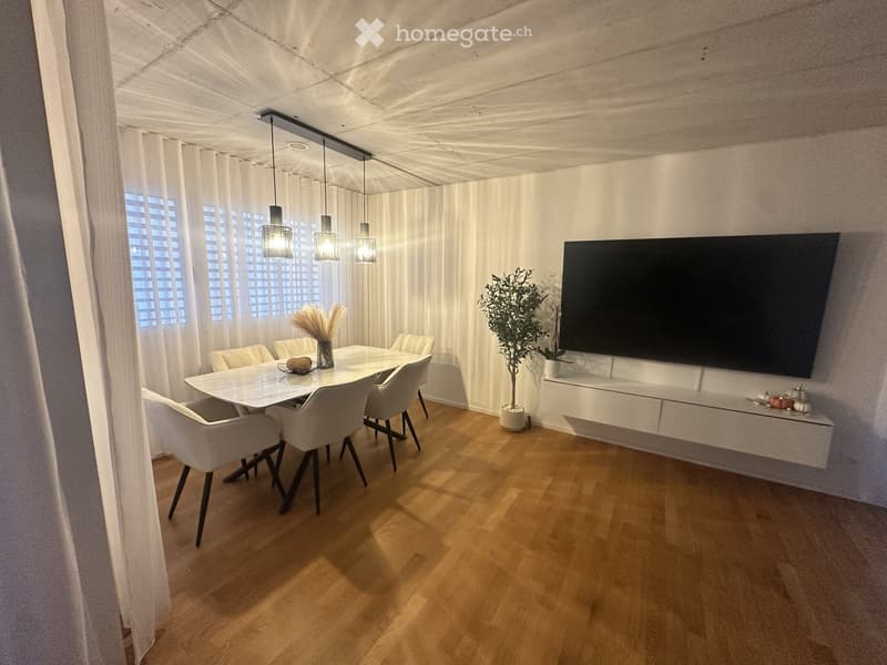 Modern dining room with a white dining table, six chairs, a large flat-screen TV mounted on the wall, pendant lights, and a potted plant. The room features wooden flooring and large windows with white curtains.