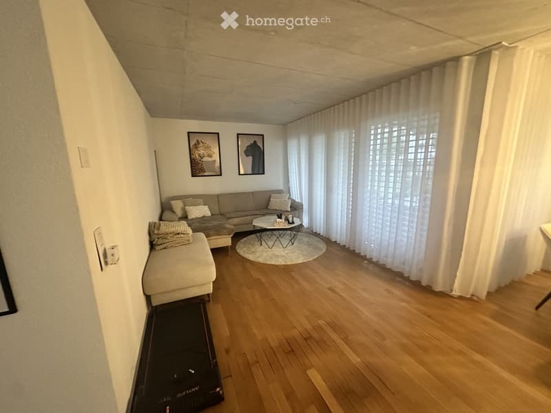 Modern living room with light wooden flooring, a grey sectional sofa, a round coffee table, an ottoman, and large windows with white curtains.