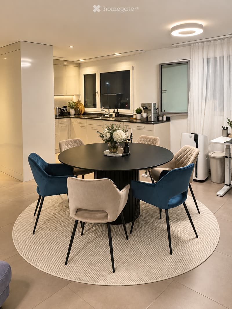 Modern dining area with a round black table surrounded by six chairs, including blue and beige upholstery. A flower vase with white flowers is placed at the center of the table. The area is adjacent to a sleek kitchen with white cabinets, a sink, and vari