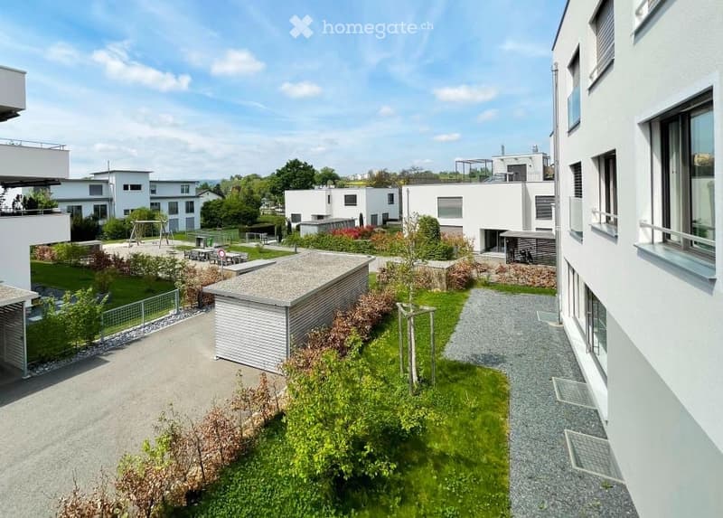 Modern residential complex with white buildings, green lawns, small trees, and paved walkways.