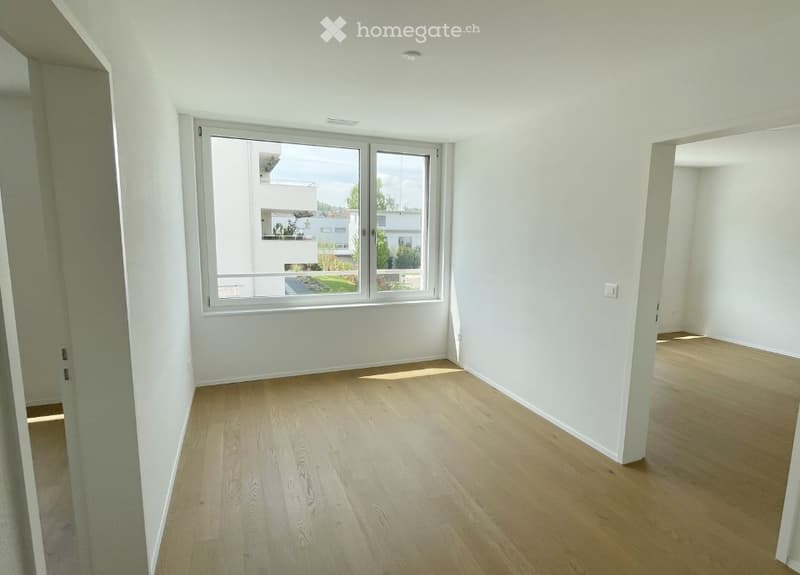 Empty room with light wooden flooring, white walls, and large windows offering a view of the outside area.