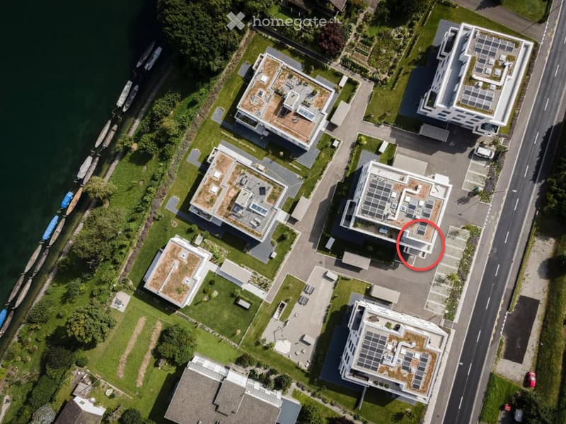 Aerial view of a residential area with multiple modern houses, green lawns, and a waterfront with boats. The houses feature flat roofs and solar panels.