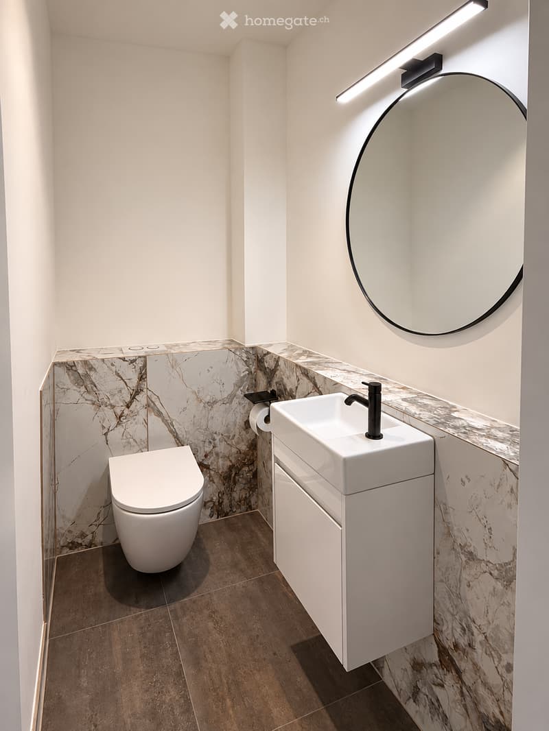 Modern bathroom with a white toilet, white sink with a black faucet, a large round mirror, and a marble backsplash.