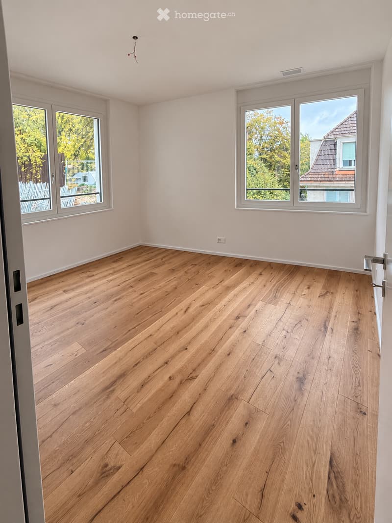 Empty room with light wooden flooring, white walls, and large windows offering a view of greenery and neighboring buildings.