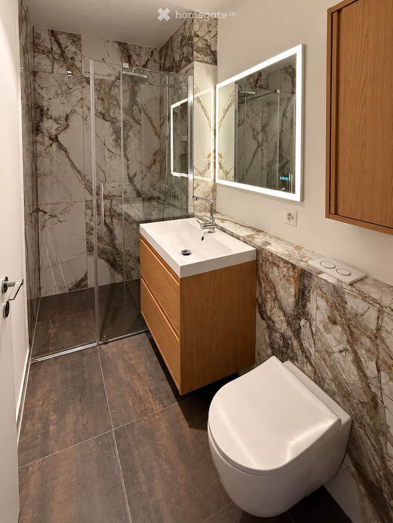 Modern bathroom with marble walls, a sleek sink with wooden cabinet, a toilet, a large mirror with LED lighting, and a glass-enclosed shower area.