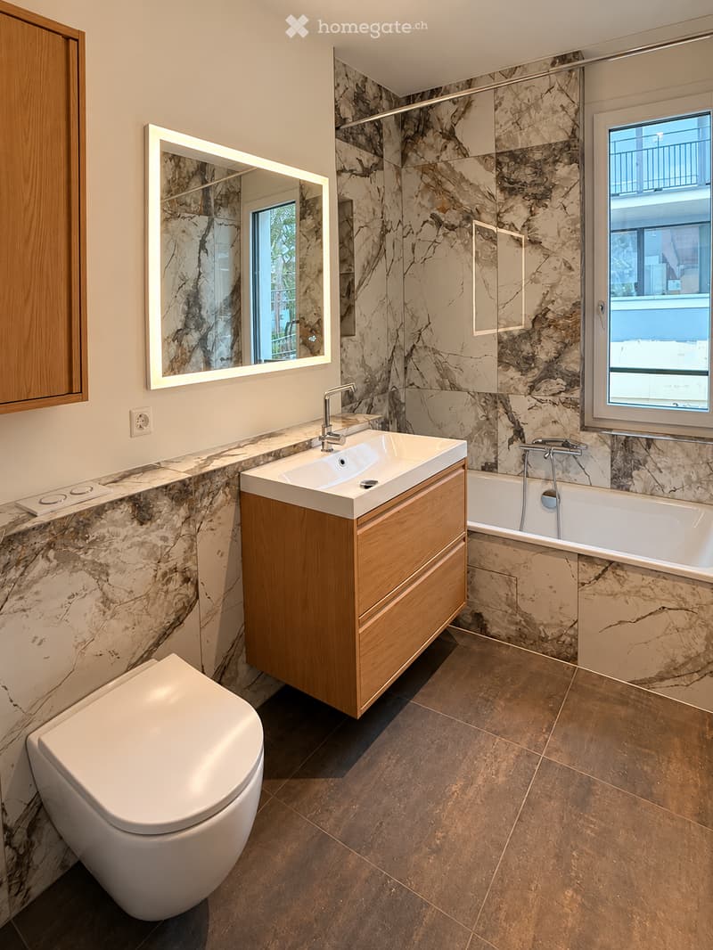 Modern bathroom with marble walls and floors, featuring a wooden vanity with a sink, a toilet, a bathtub, and a large mirror with built-in lighting.