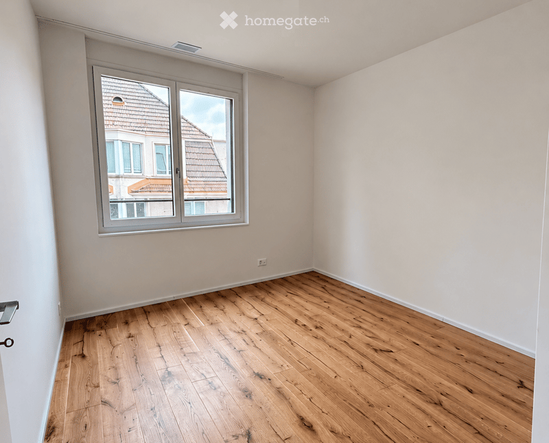 Empty room with wooden flooring and large window showing a view of neighboring buildings.