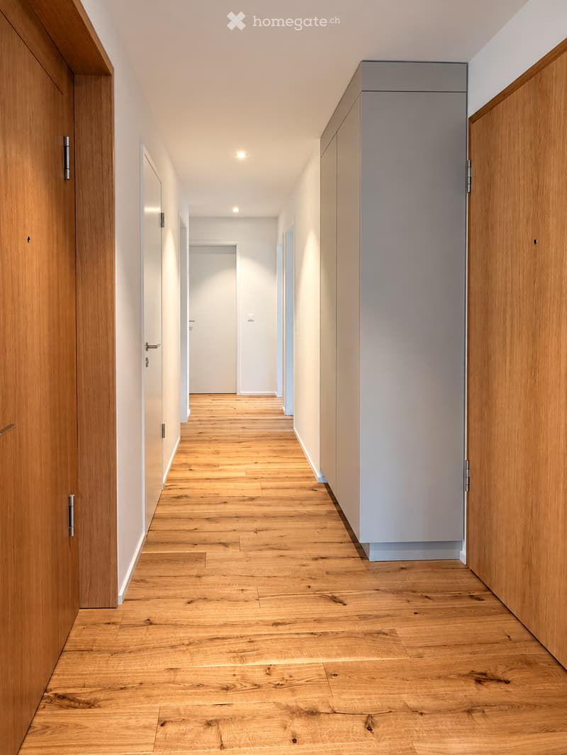 Modern hallway with wooden flooring, white walls, and built-in wooden cabinetry on one side. Features recessed lighting and multiple doors leading to other rooms.
