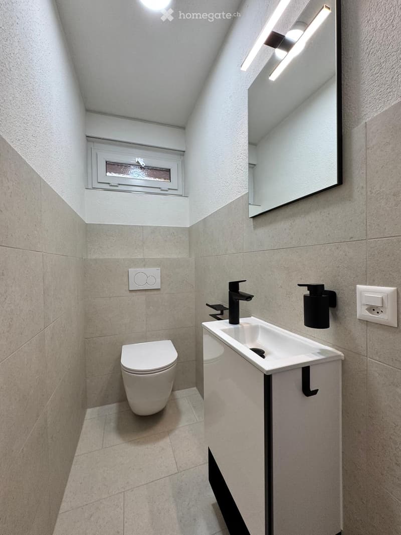 Modern bathroom with a toilet, sink, and mirror. Features a sleek black faucet and a built-in soap dispenser. The room has a sloped ceiling and a small window for ventilation.