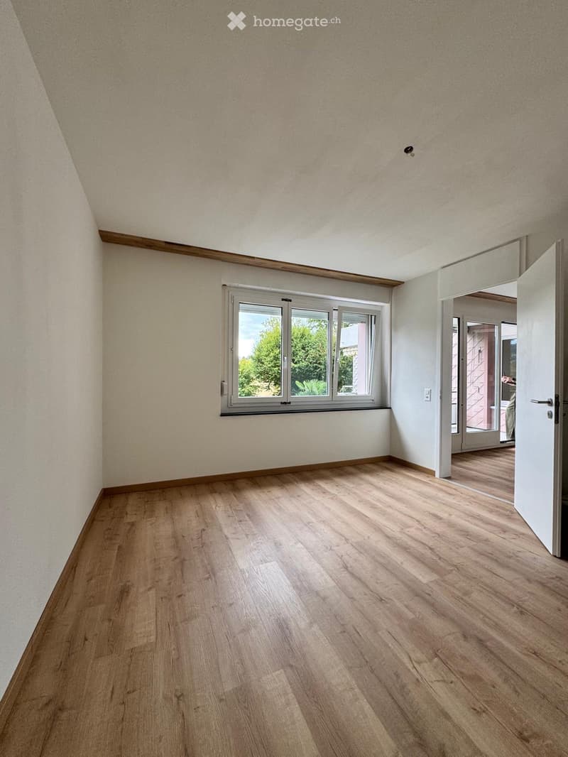 Empty room with wooden flooring, white walls, and a large window with a view of greenery. The room has a slanted ceiling and an open doorway leading to another room.