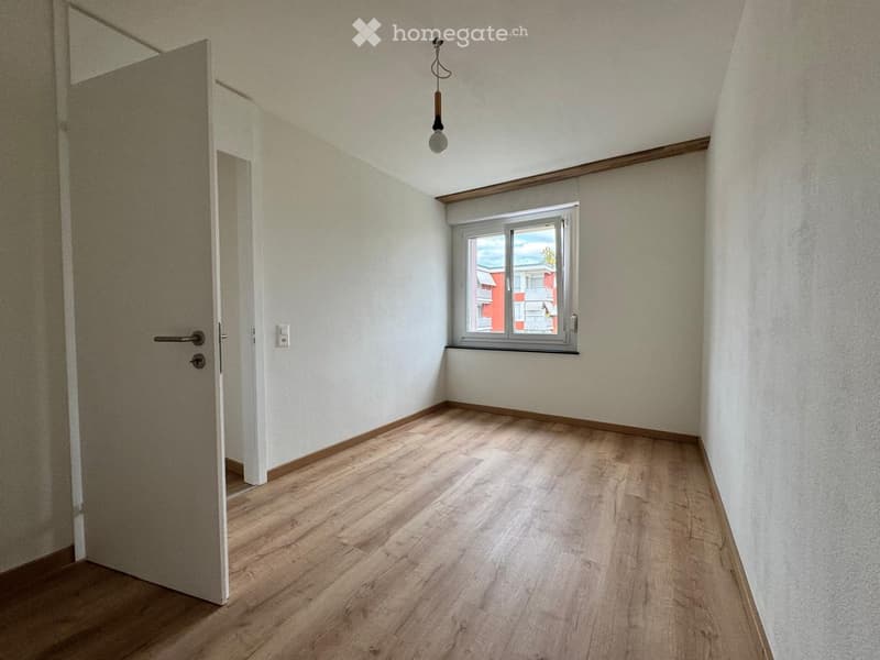 Empty room with light wooden flooring, white walls, a single window, and a door leading to another space.