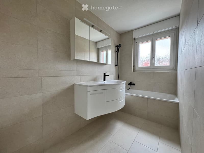 Modern bathroom with a white sink, mirror, bathtub, and showerhead. The walls and floor are tiled in a light beige color.